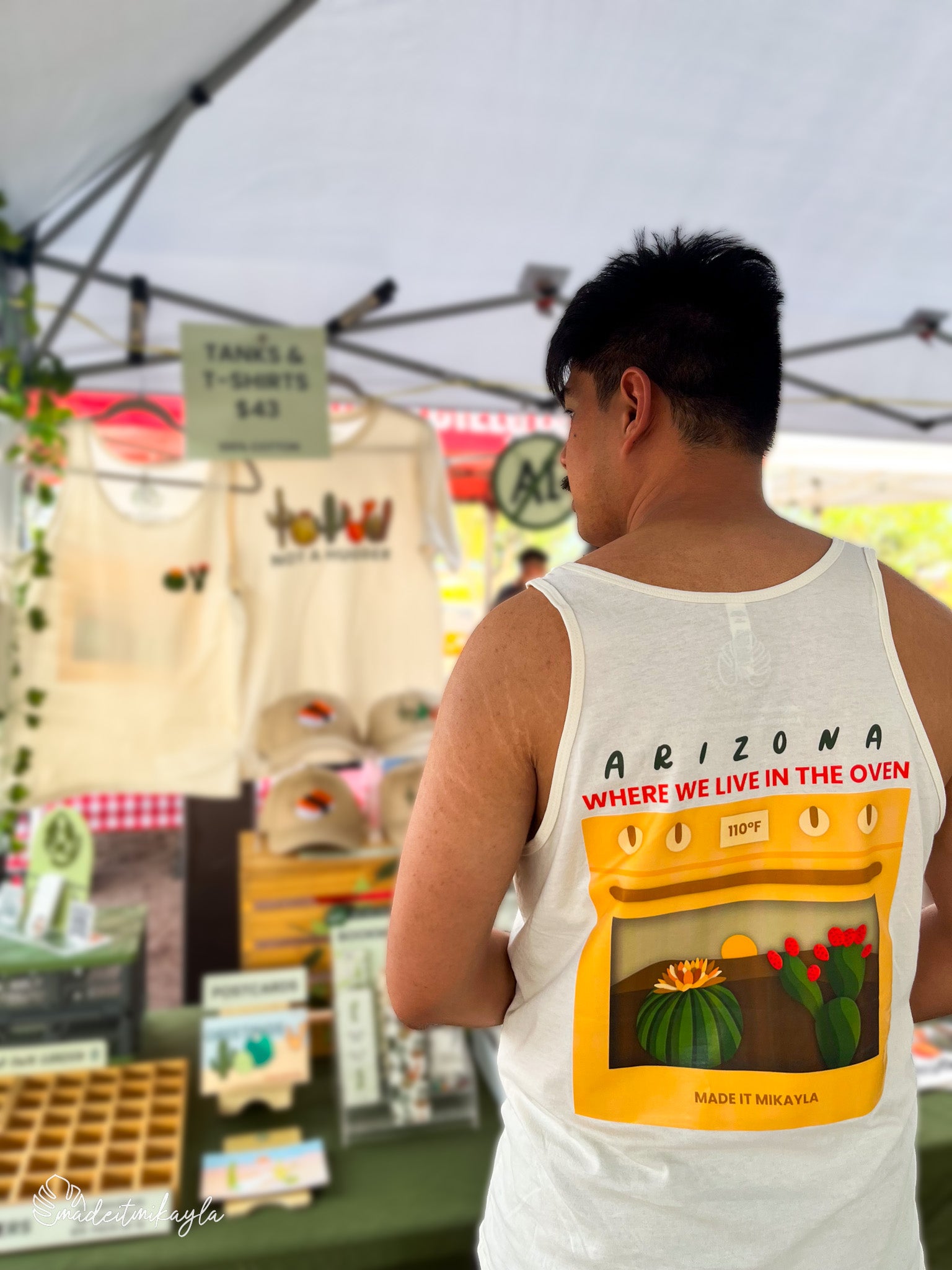 Booth bae wearing Arizona Oven tank top with back graphic of an oven with cacti in it, and text 'Arizona, where we live in the oven' at an outdoor market.