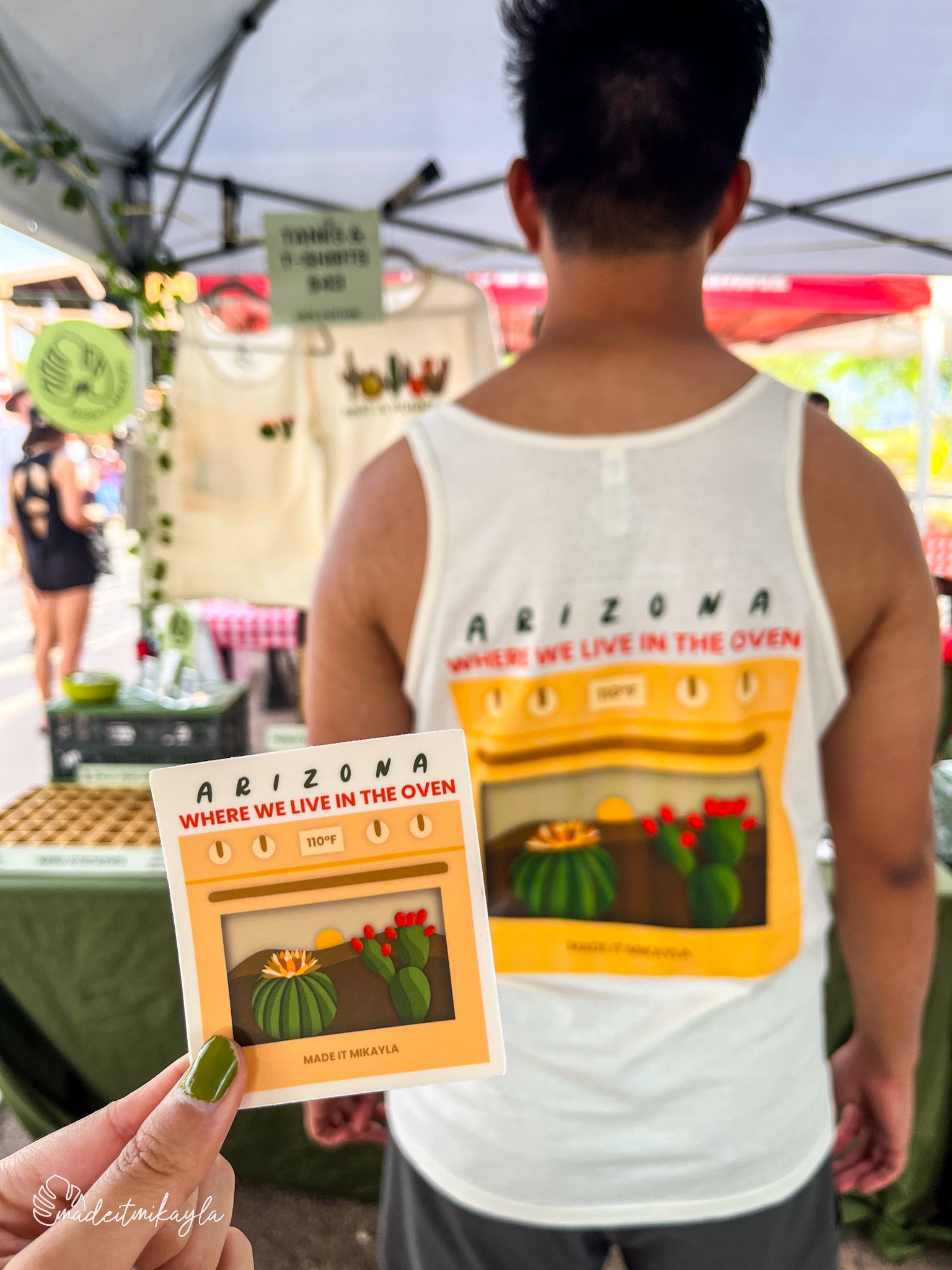 Booth bae wearing Arizona Oven tank top with cactus design, with a hand holding matching Arizona Oven sticker at an outdoor market.