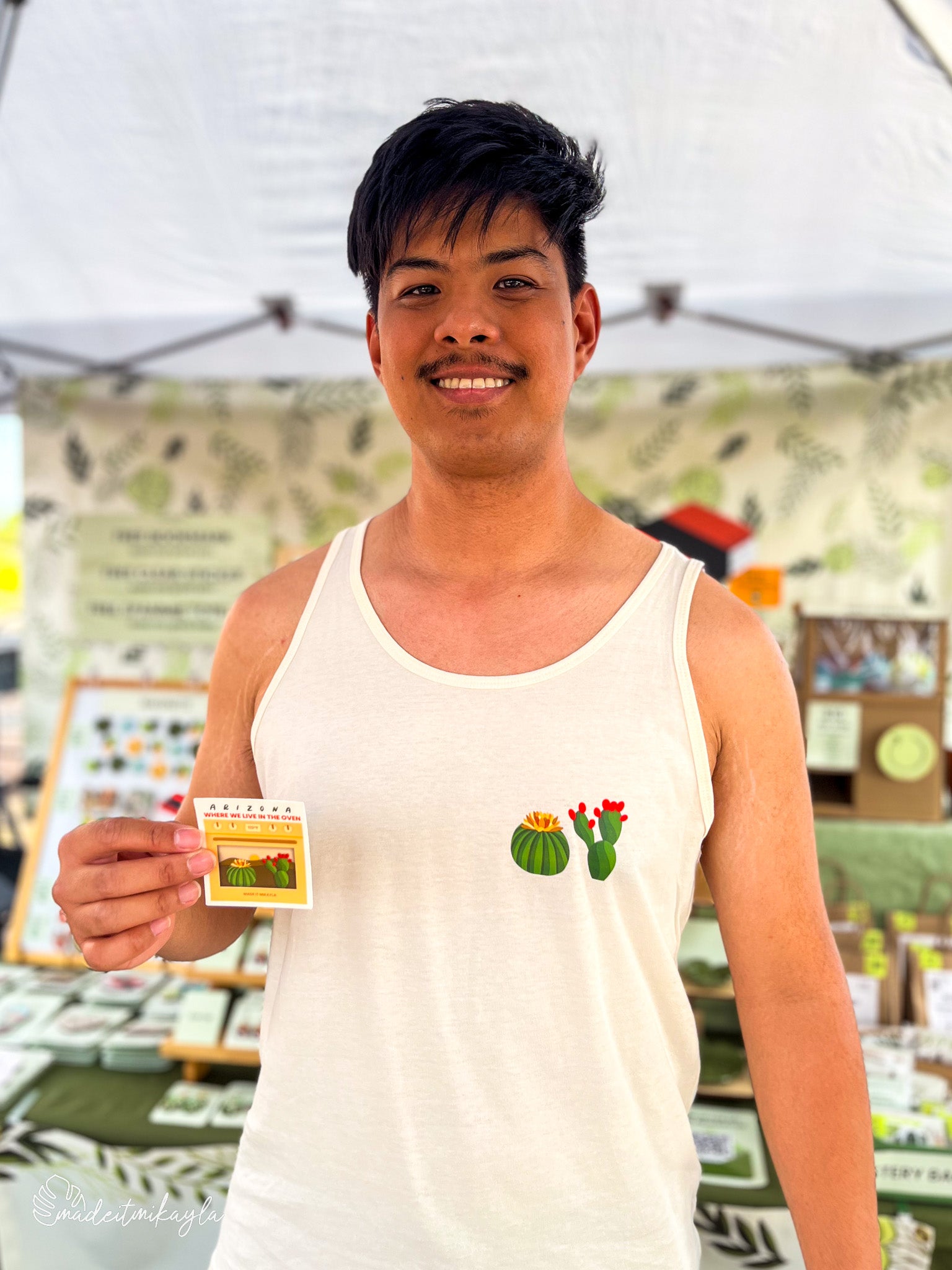 Booth bae holding Arizona Oven sticker while wearing matching Arizona Oven tank top with cactus design at an outdoor market.