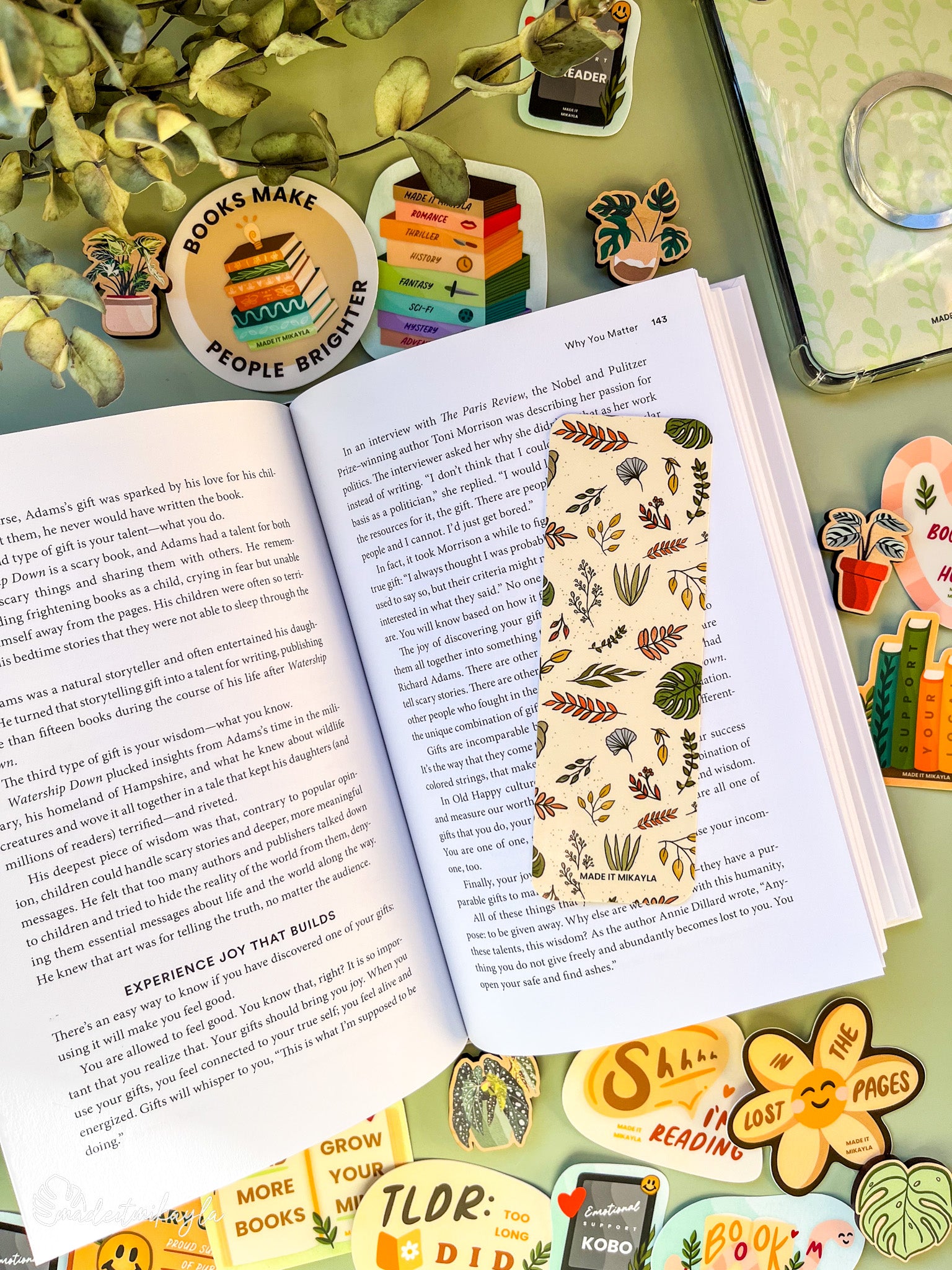 Open book with a bookmark featuring leaves on a yellow background, surrounded by bookish stickers and decorations on a green surface.