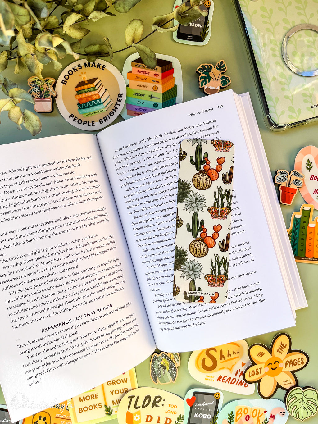 Open book with a bookmark featuring cacti and succulents on a green background, surrounded by bookish stickers and decorations on a green surface.