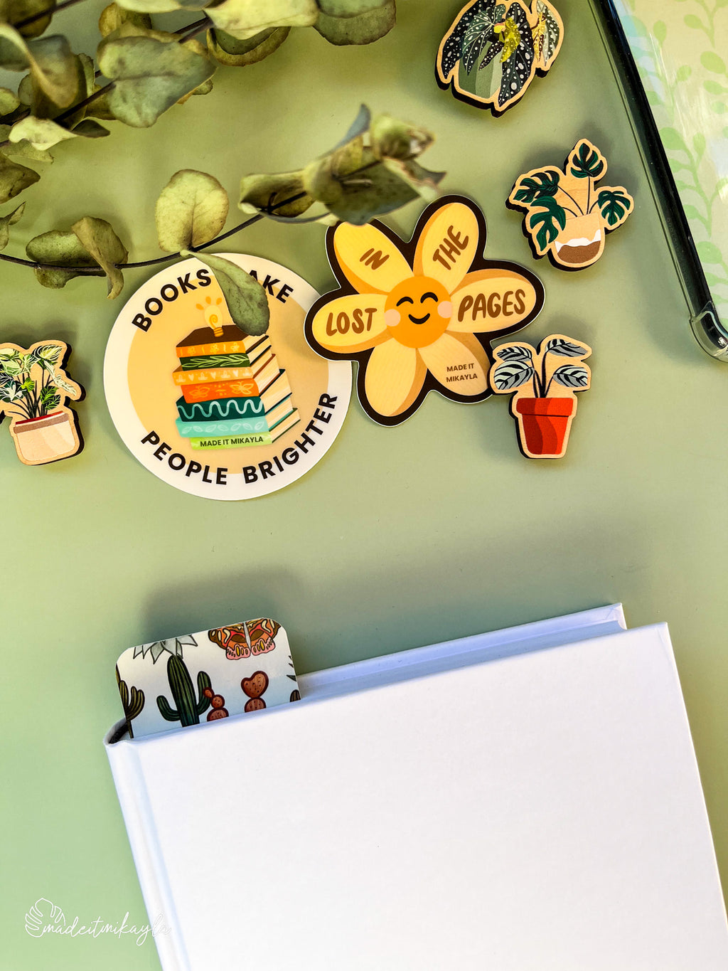 Closed book with a bookmark sticking out the top, featuring cacti, surrounded by bookish stickers and decorations on a green surface.