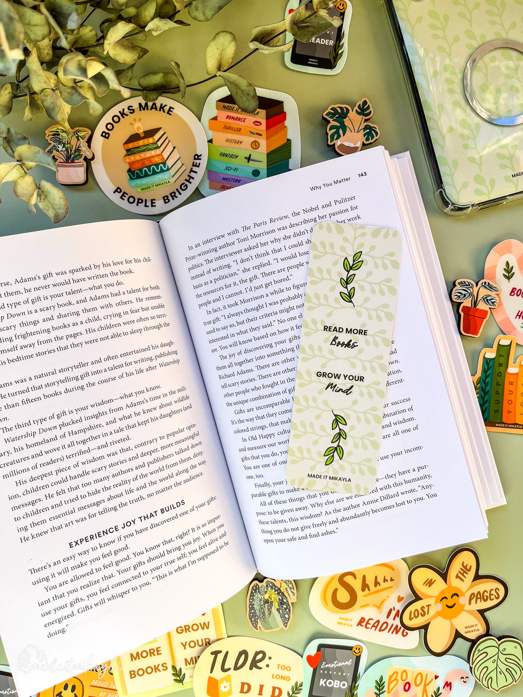 Open book with a bookmark featuring green leaves and the quote "Read more books grow your mind," surrounded by bookish stickers and decorations on a green surface.
