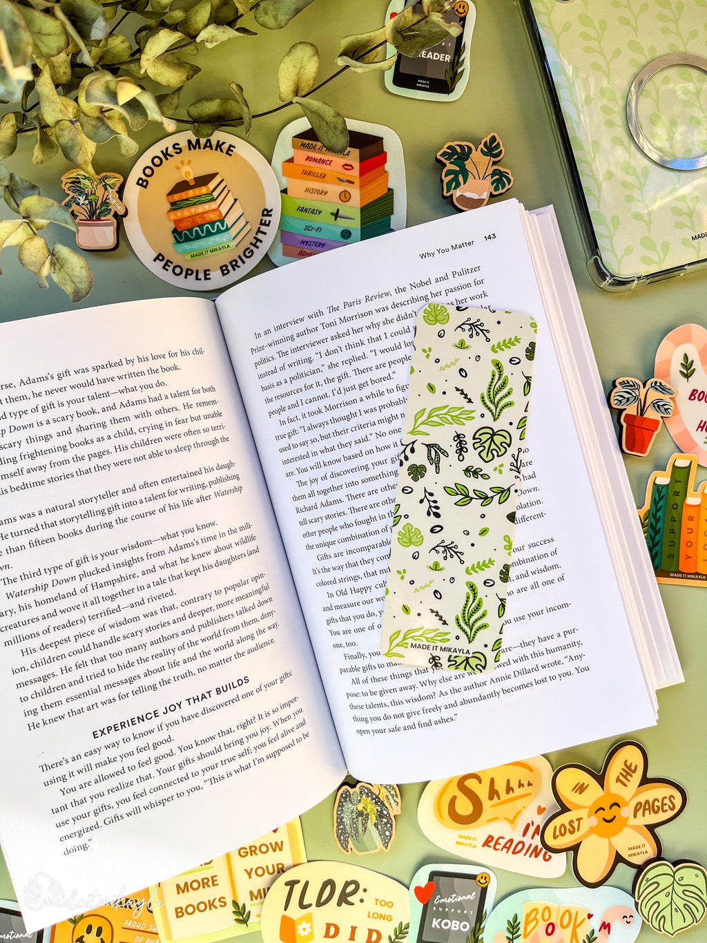Open book with a bookmark featuring green leaves on a green background, surrounded by bookish stickers and decorations on a green surface.