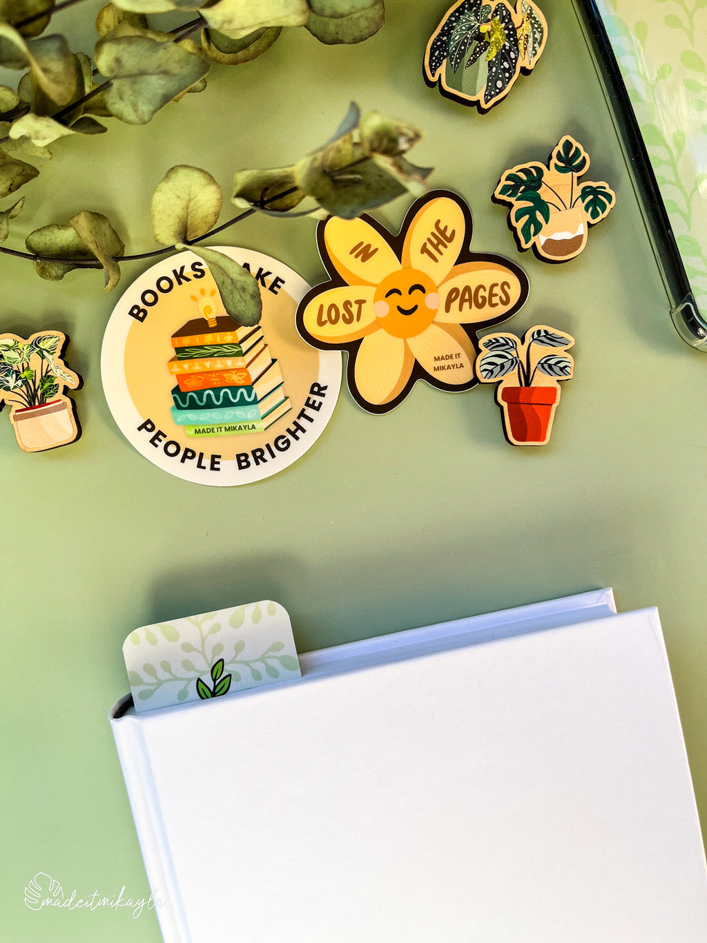 Closed book with a bookmark sticking out the top, featuring green leaves, surrounded by bookish stickers and decorations on a green surface.