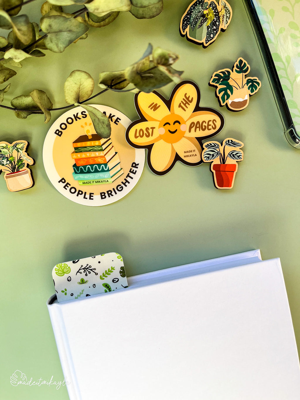 Closed book with a bookmark sticking out the top, featuring green leaves, surrounded by bookish stickers and decorations on a green surface.