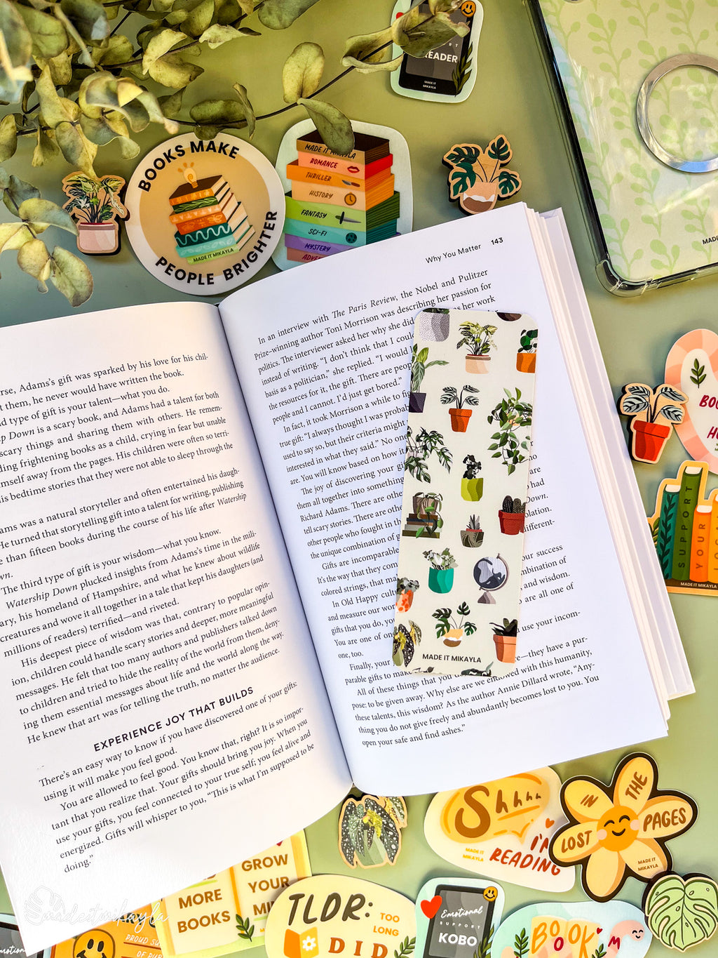 Open book with a bookmark featuring potted plants scattered on a green background, surrounded by bookish stickers and decorations on a green surface.
