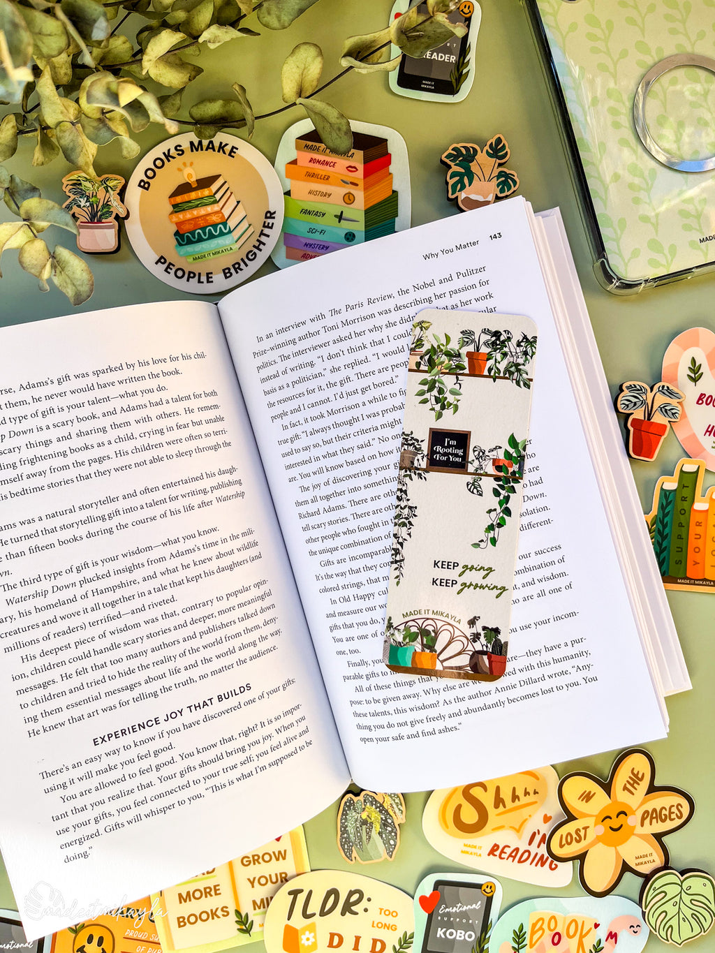 Open book with a bookmark featuring potted plants on shelves and the quote "keep going keep growing" on a green background, surrounded by bookish stickers and decorations on a green surface.