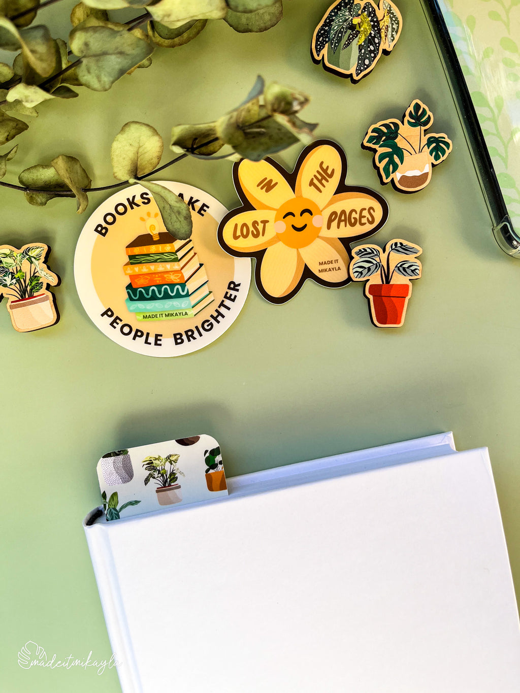 Closed book with a bookmark sticking out the top, featuring potted plants, surrounded by bookish stickers and decorations on a green surface.