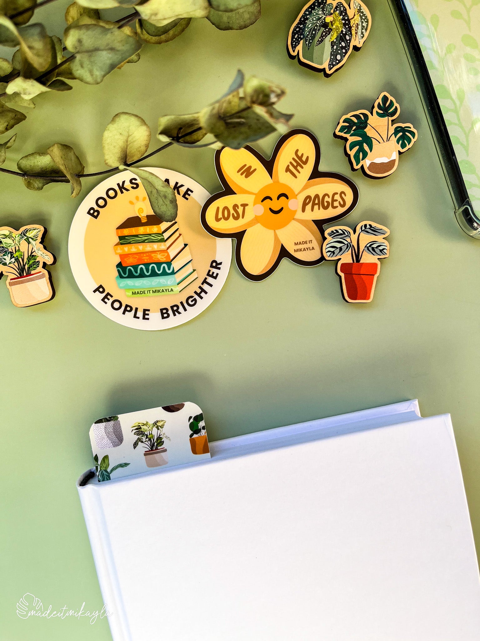 Closed book with a bookmark sticking out the top, featuring potted plants, surrounded by bookish stickers and decorations on a green surface.