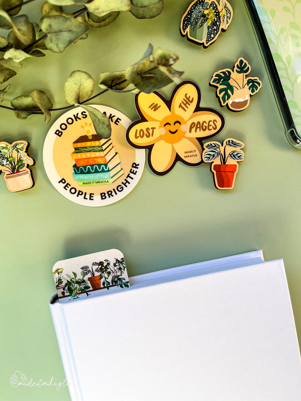 Closed book with a bookmark sticking out the top, featuring potted plants on a shelf, surrounded by bookish stickers and decorations on a green surface.