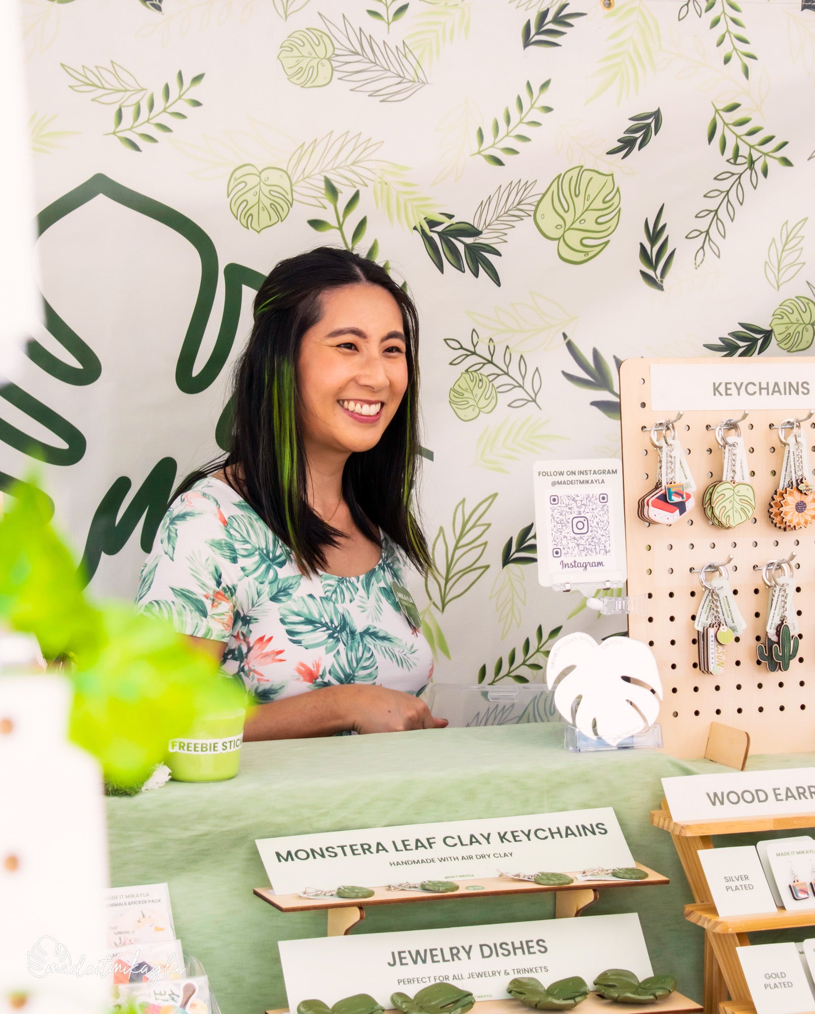 Woman standing behind a display of handmade items with a leaf-patterned wall in the background