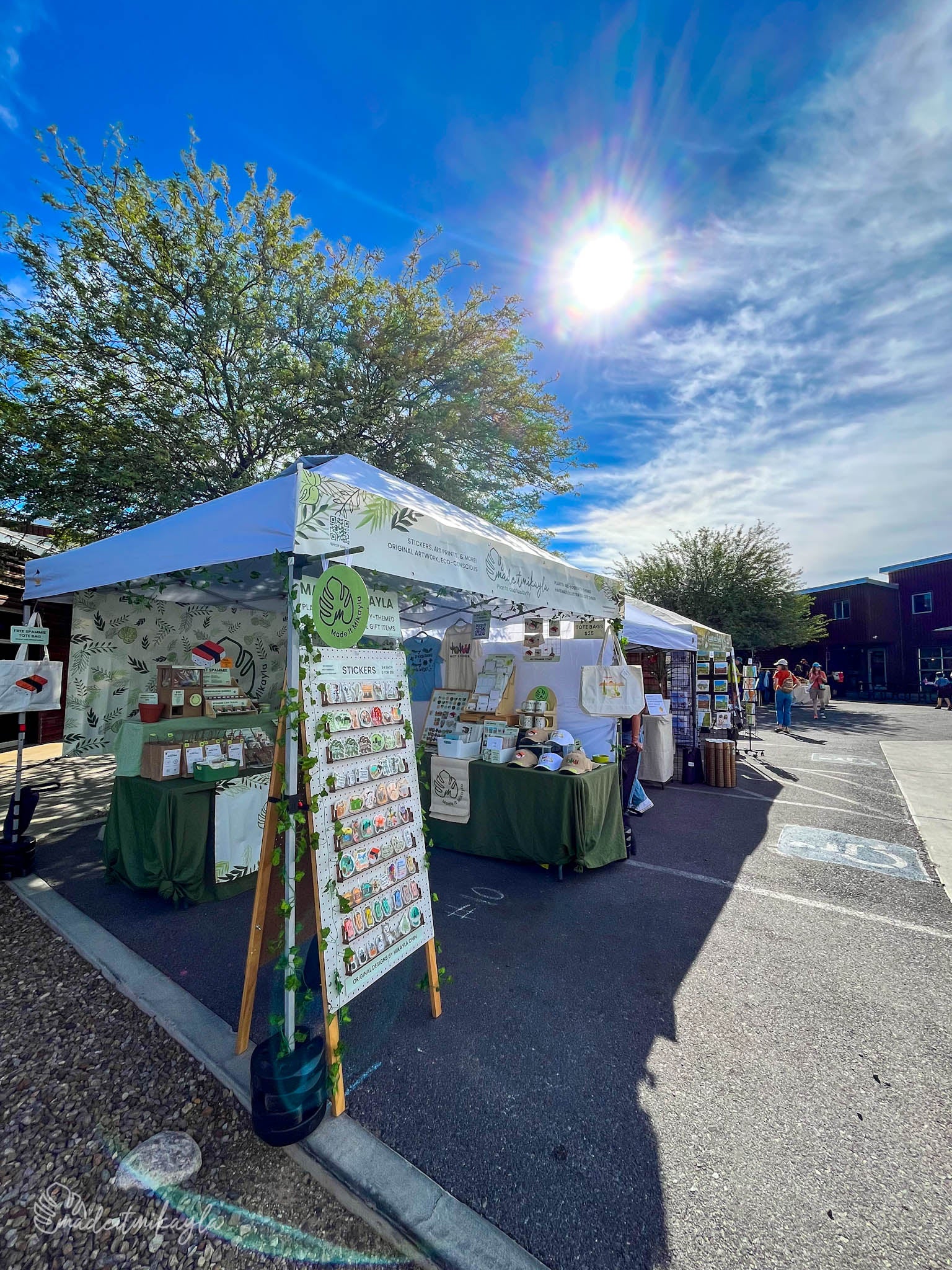 Outdoor market stall with MadeItMikayla products under a white tent on a sunny day.