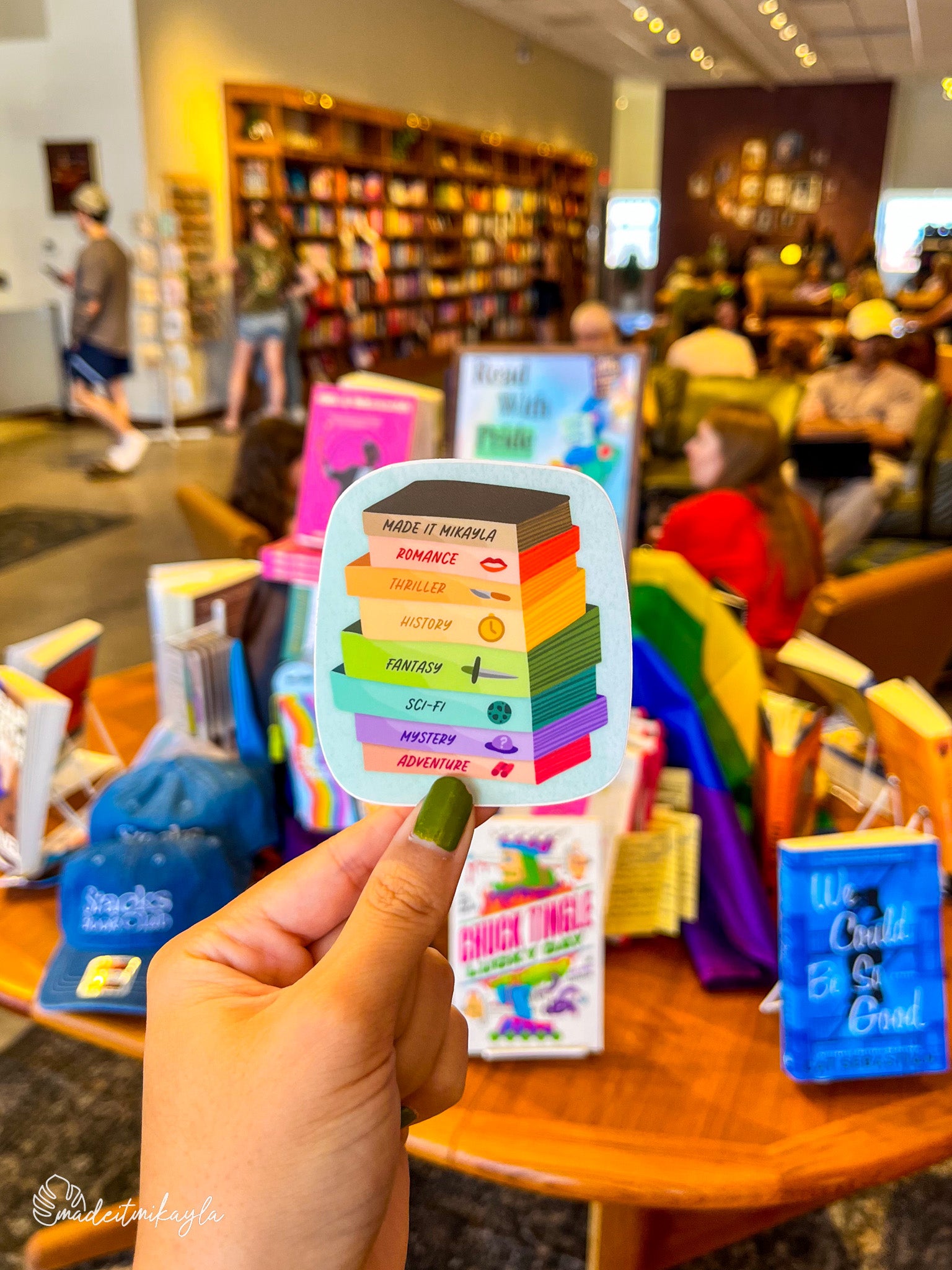 Hand holding colorful 'Book Stack Genres' sticker in front of a bookstore interior with people browsing books.
