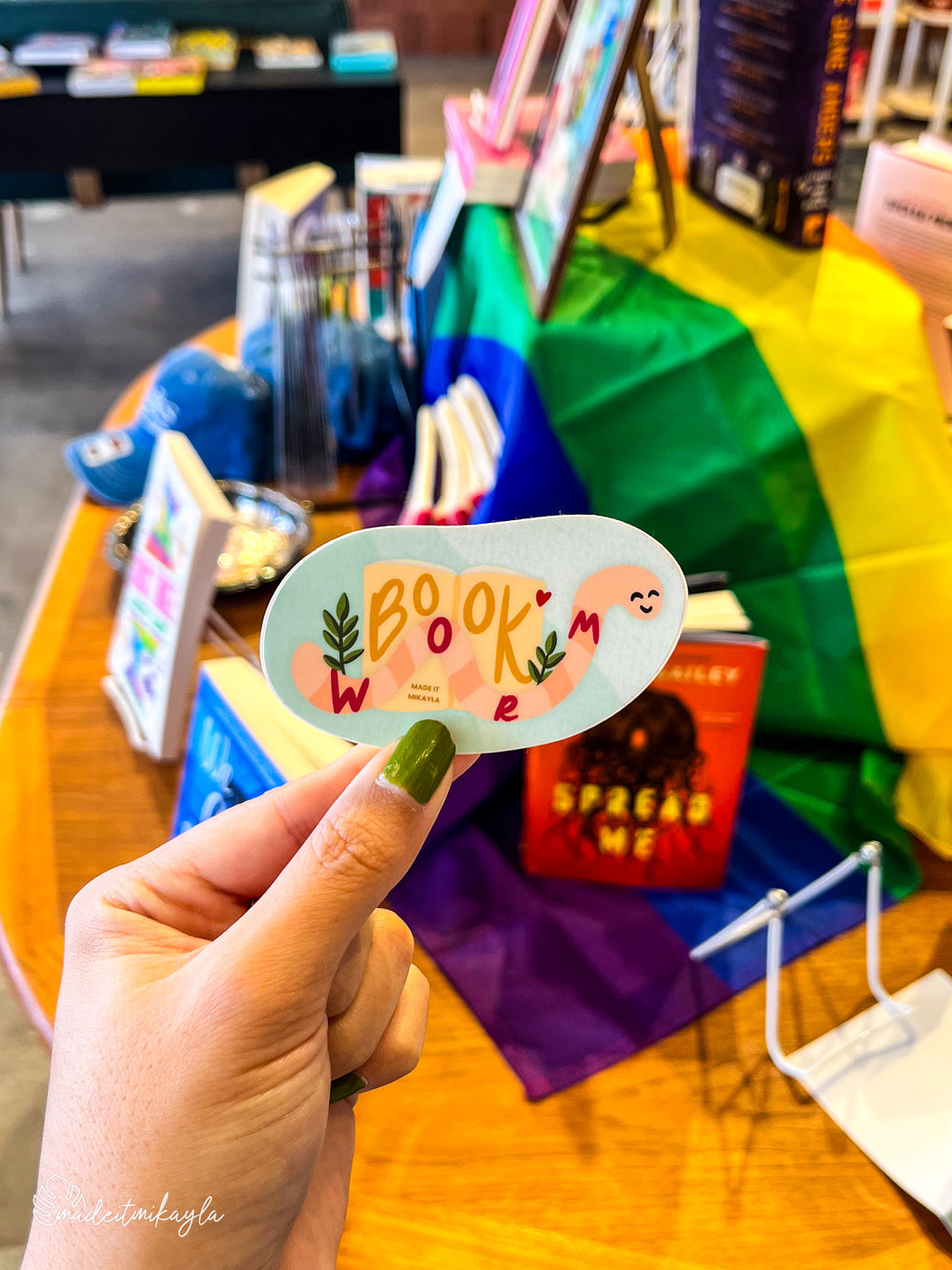 Hand holding a 'Book Worm' sticker with a colorful background of books and a rainbow flag.