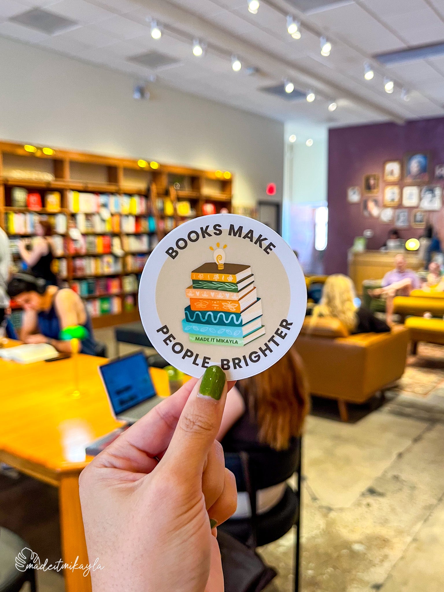 Hand holding a 'Books Make People Brighter' sticker in a bookstore.
