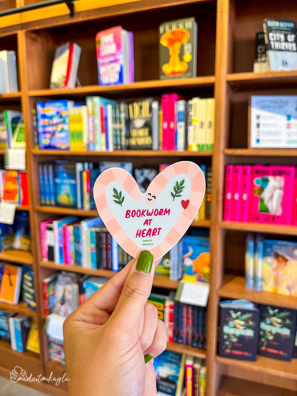 Hand holding a heart-shaped 'Bookworm at Heart' sticker in front of a bookshelf filled with books.