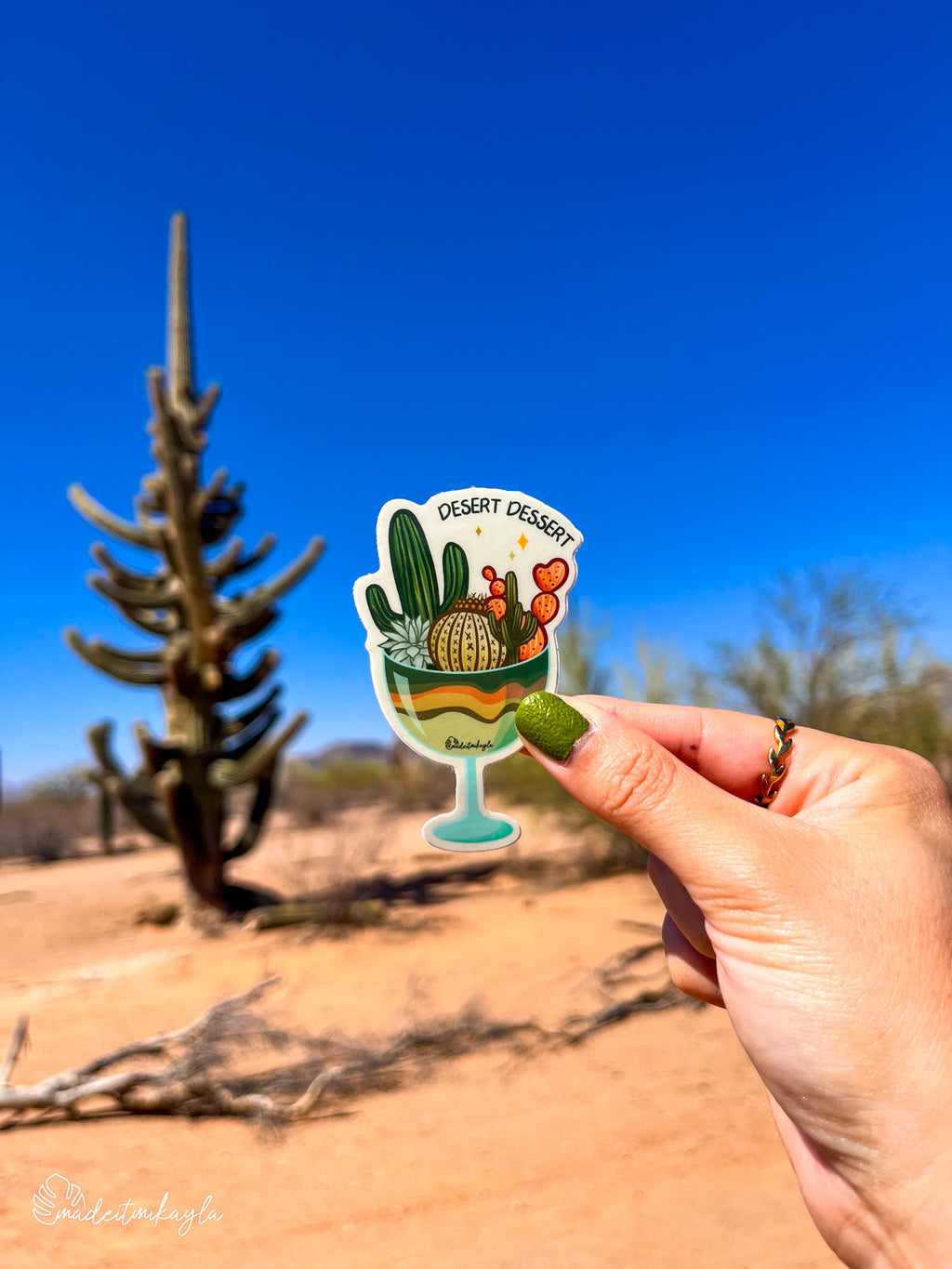 Hand holding a desert-themed sticker with a large cactus against a clear blue sky and desert landscape.