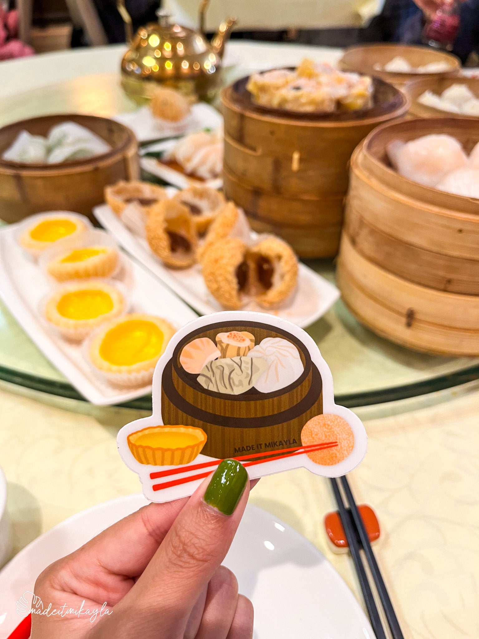 Dim Sum Sticker held by a hand with dim sum in the background.