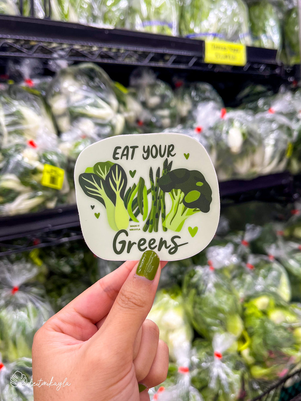Hand holding a 'Eat Your Greens' sticker in front of a green vegetable produce section.