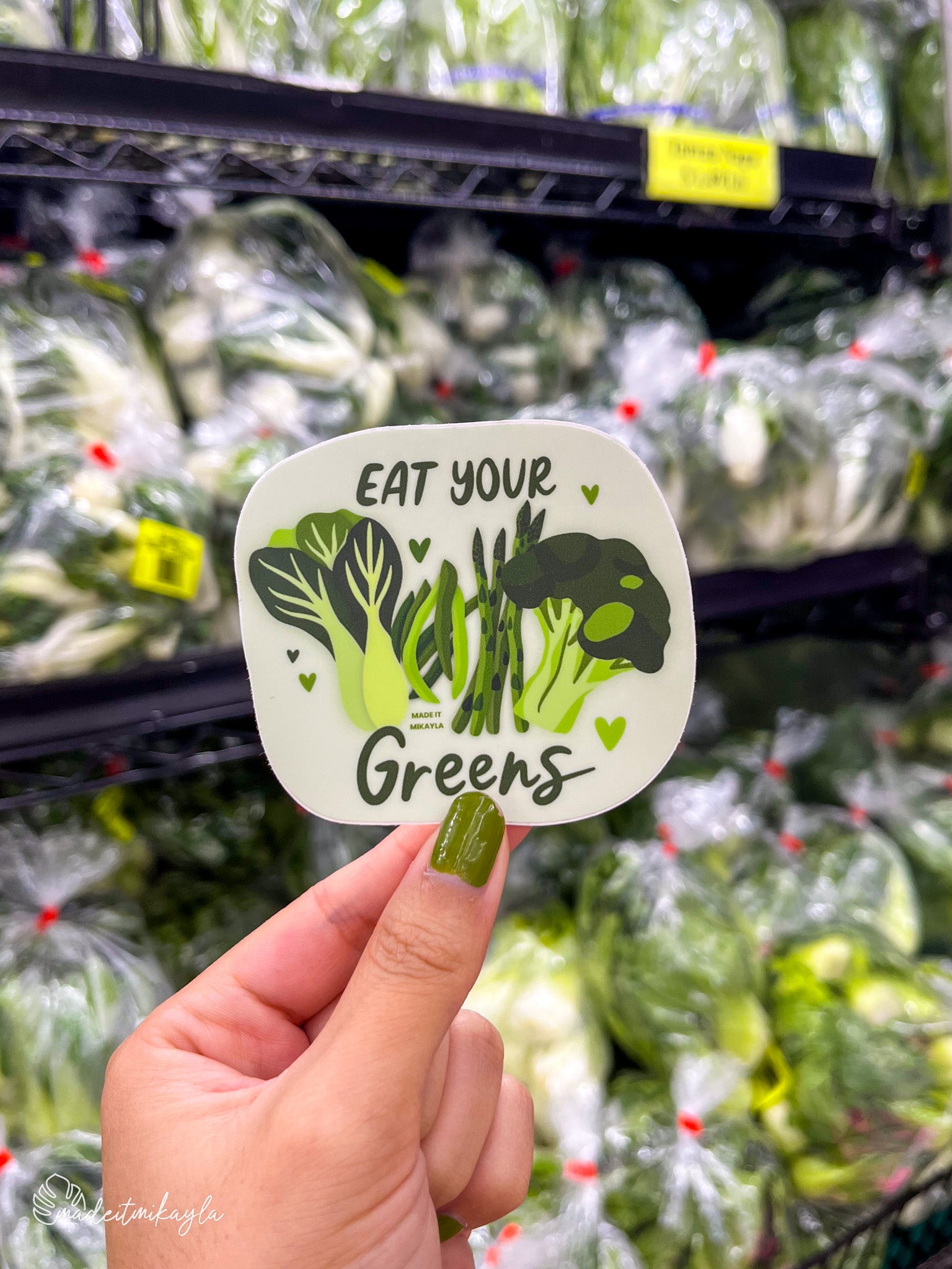 Hand holding a 'Eat Your Greens' sticker in front of a green vegetable produce section.