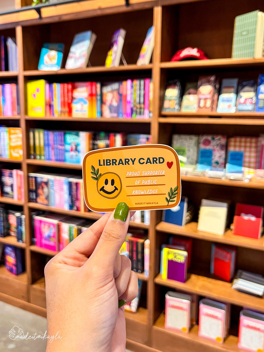 Hand holding a smiley face 'Library Card' sticker in front of a bookshelf filled with colorful books.