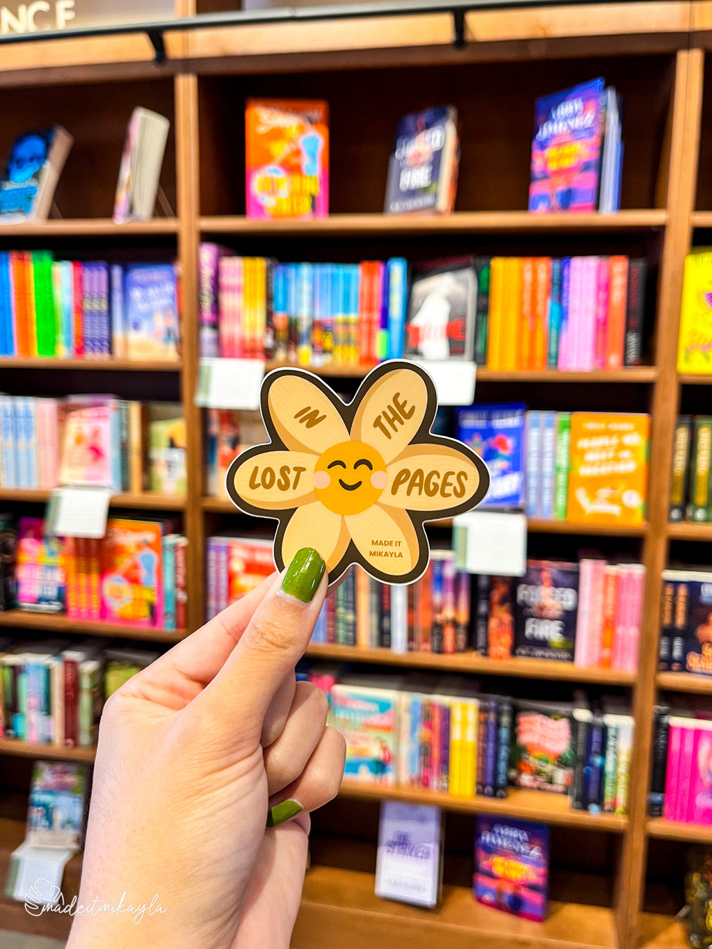 Hand holding a 'Lost In The Pages' sticker in front of a bookshelf filled with colorful books.