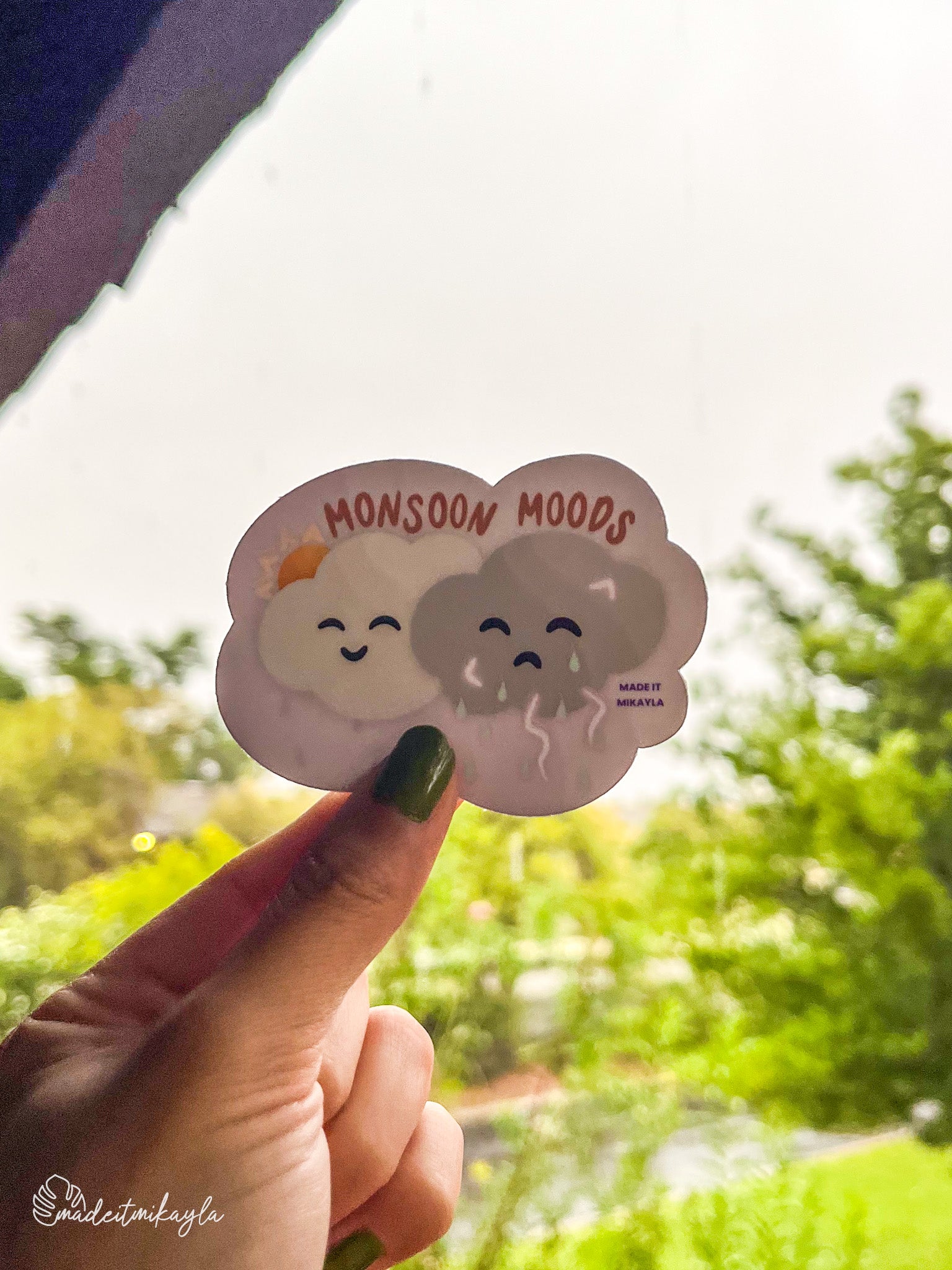 Sticker with handdrawn clouds and 'Monsoon Moods' text held by a hand against a green outdoor background.