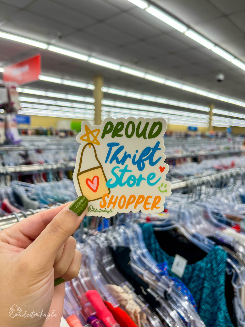 Hand holding a 'Proud Thrift Store Shopper' sticker in a clothing thrift store.