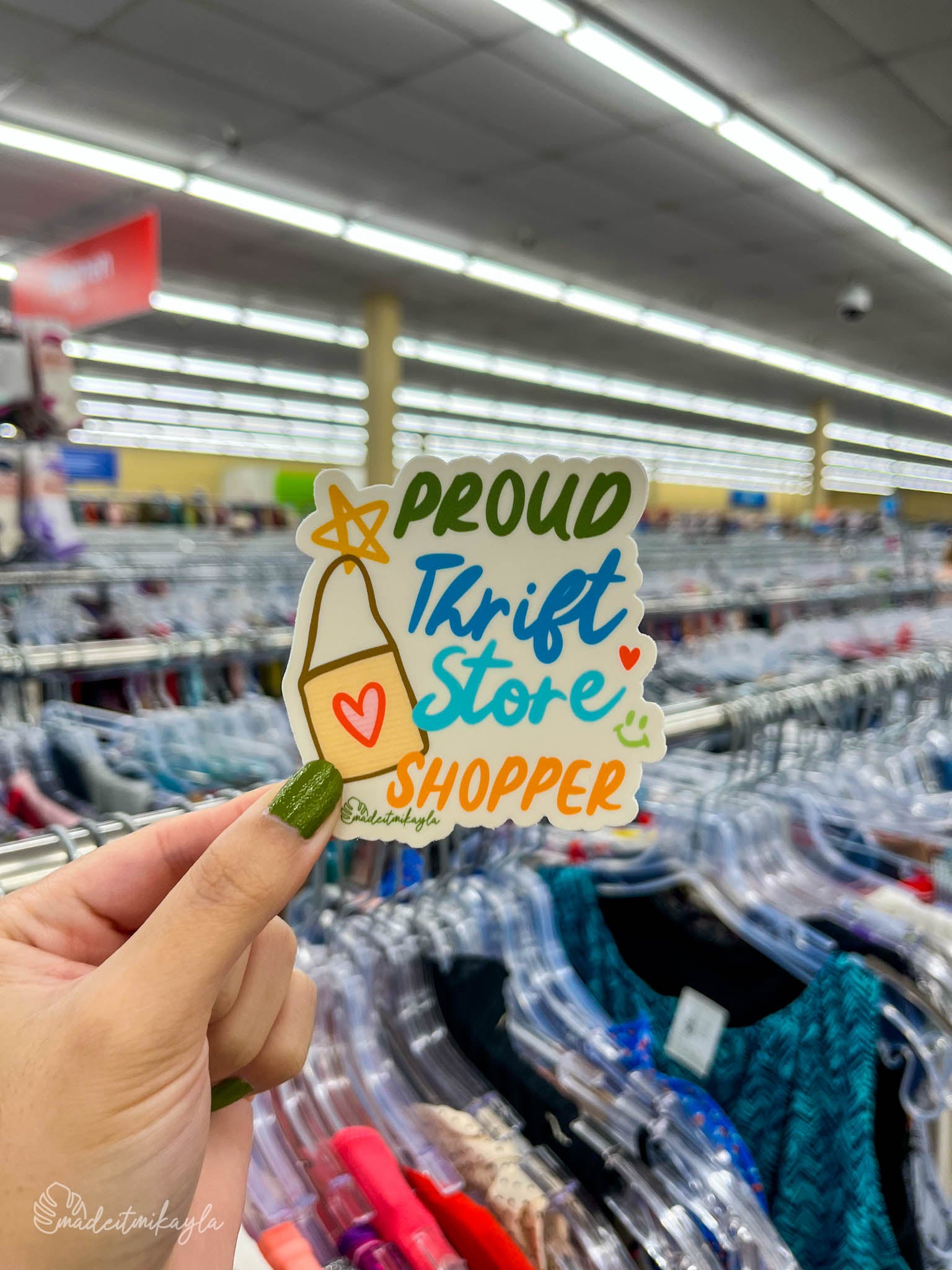 Hand holding a 'Proud Thrift Store Shopper' sticker in a clothing thrift store.