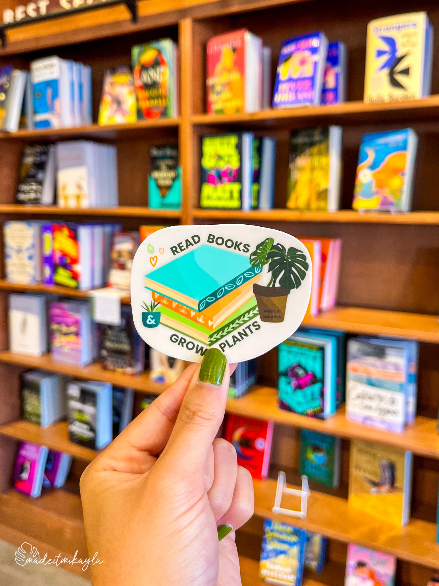 Hand holding a 'Read Books & Grow Plants' sticker in front of a bookshelf.