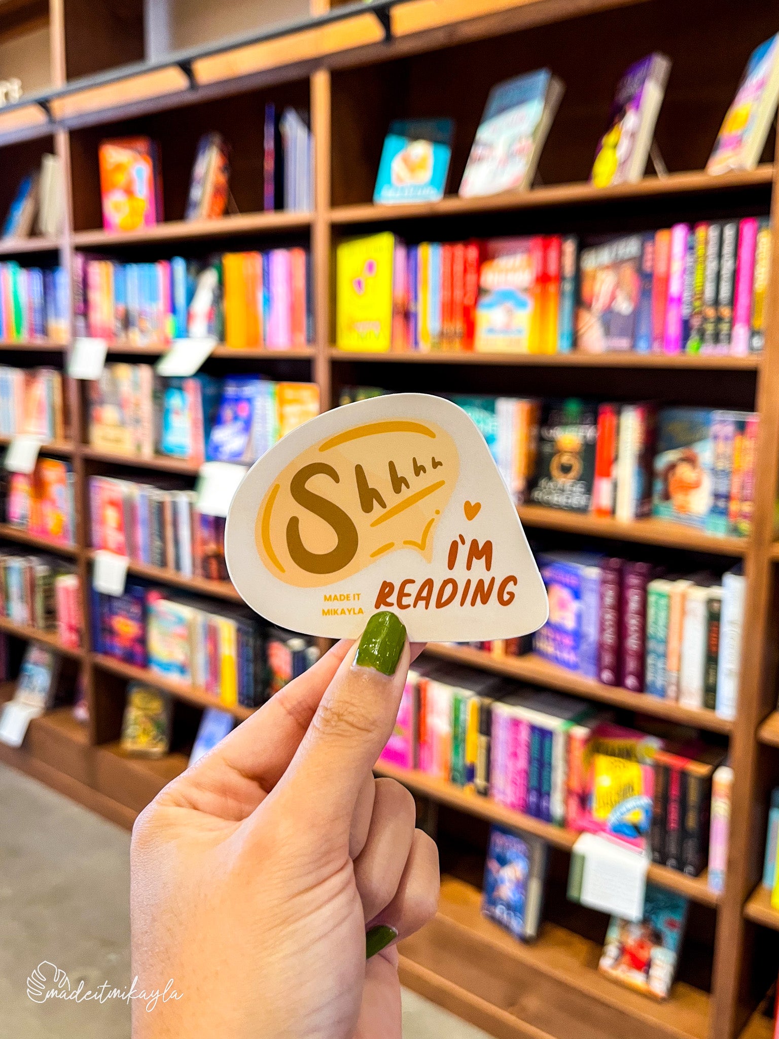 Hand holding a 'Shhhh I'm Reading' sticker in front of a bookshelf filled with colorful books.