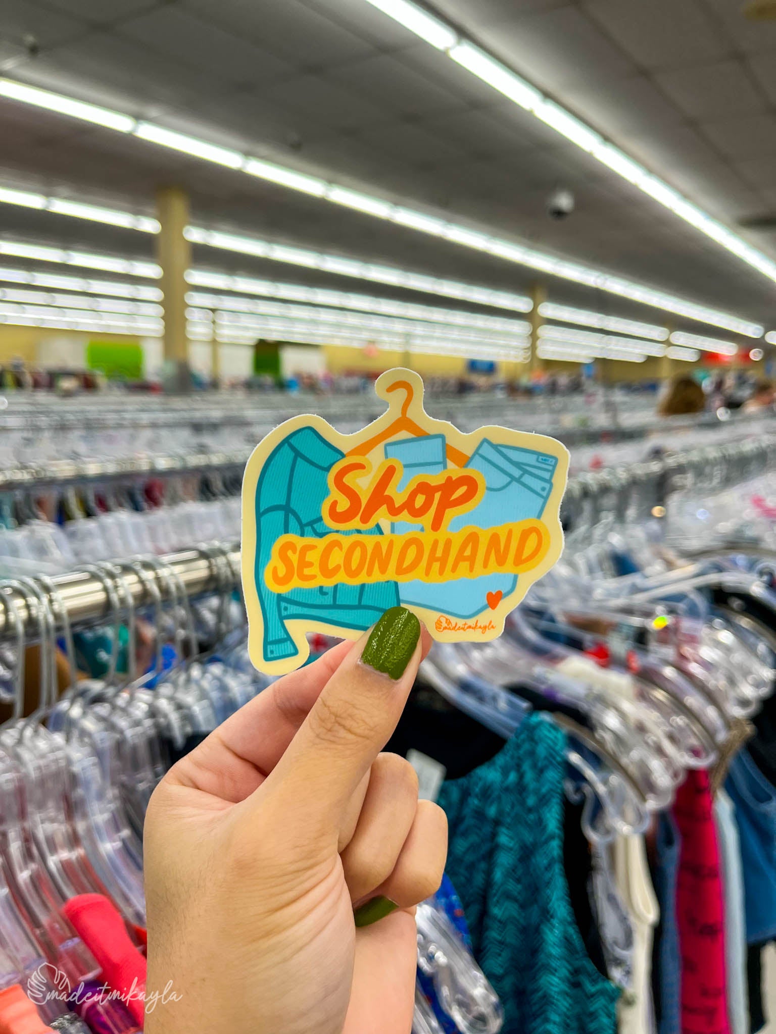 Hand holding a 'Shop Secondhand' sticker in a clothing thrift store.