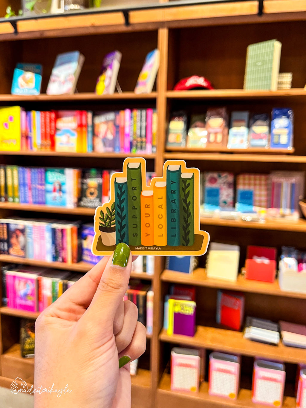 Hand holding a 'Support Your Local Library' sticker in front of a bookshelf. 