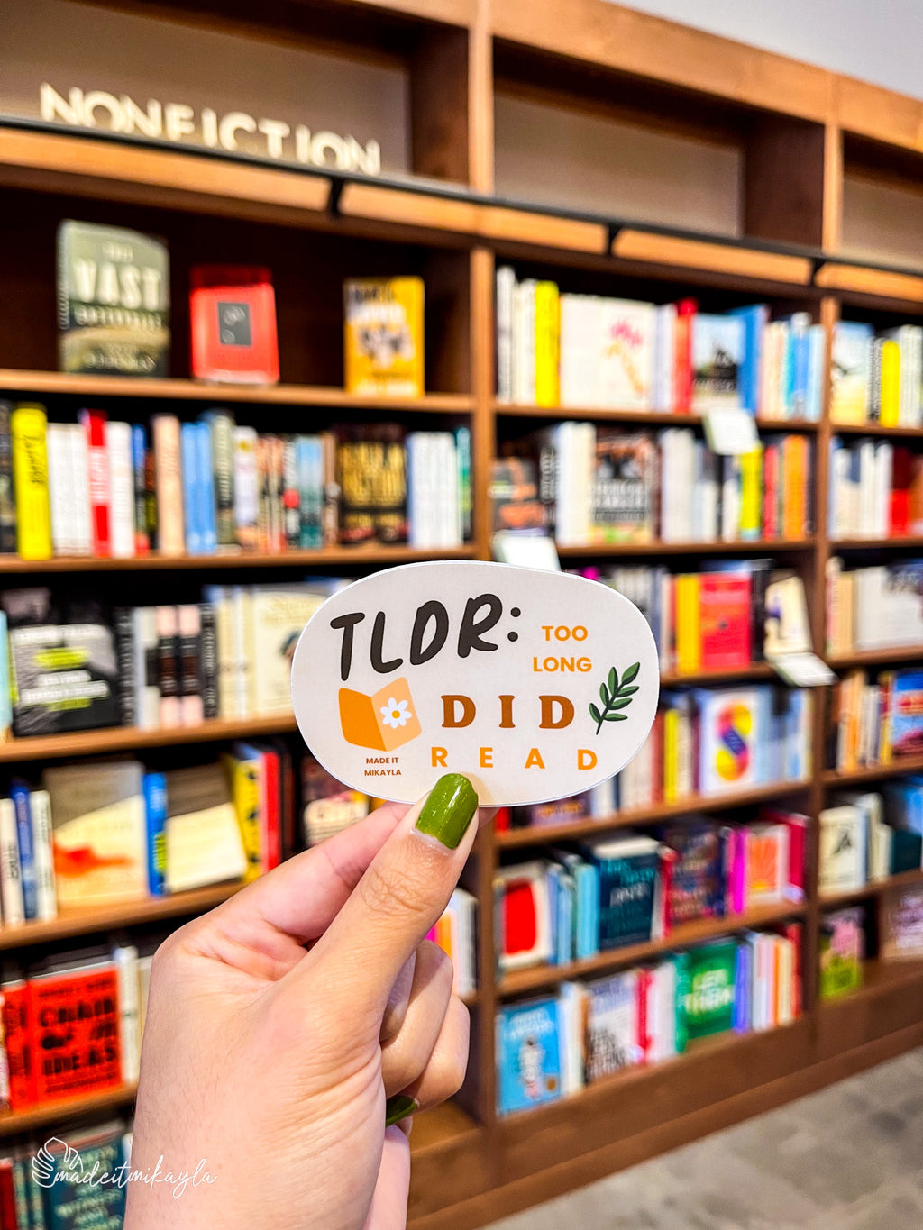 Hand holding a 'TLDR: Too Long Did Read' sticker in front of a bookshelf.