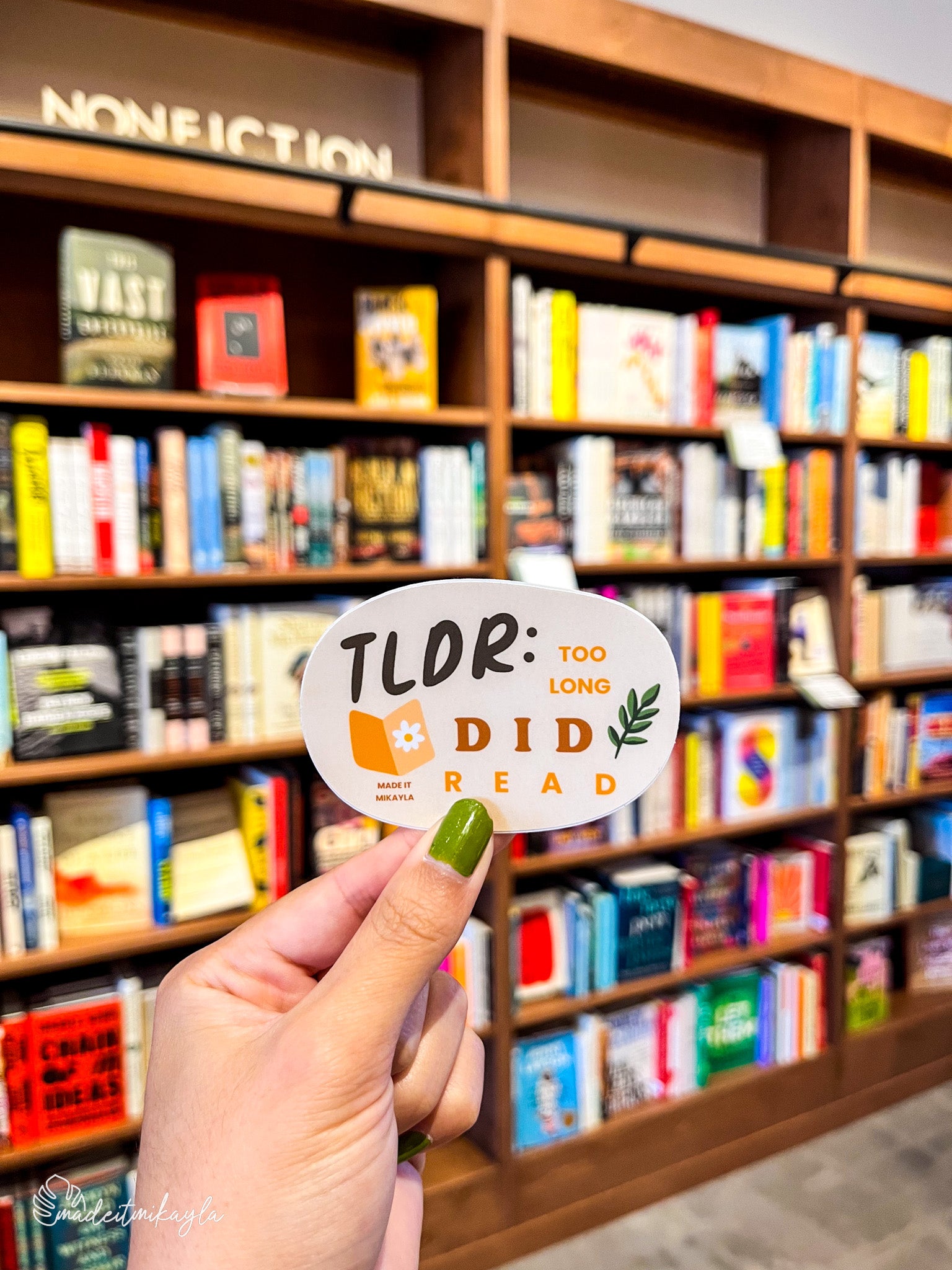 Hand holding a 'TLDR: Too Long Did Read' sticker in front of a bookshelf.