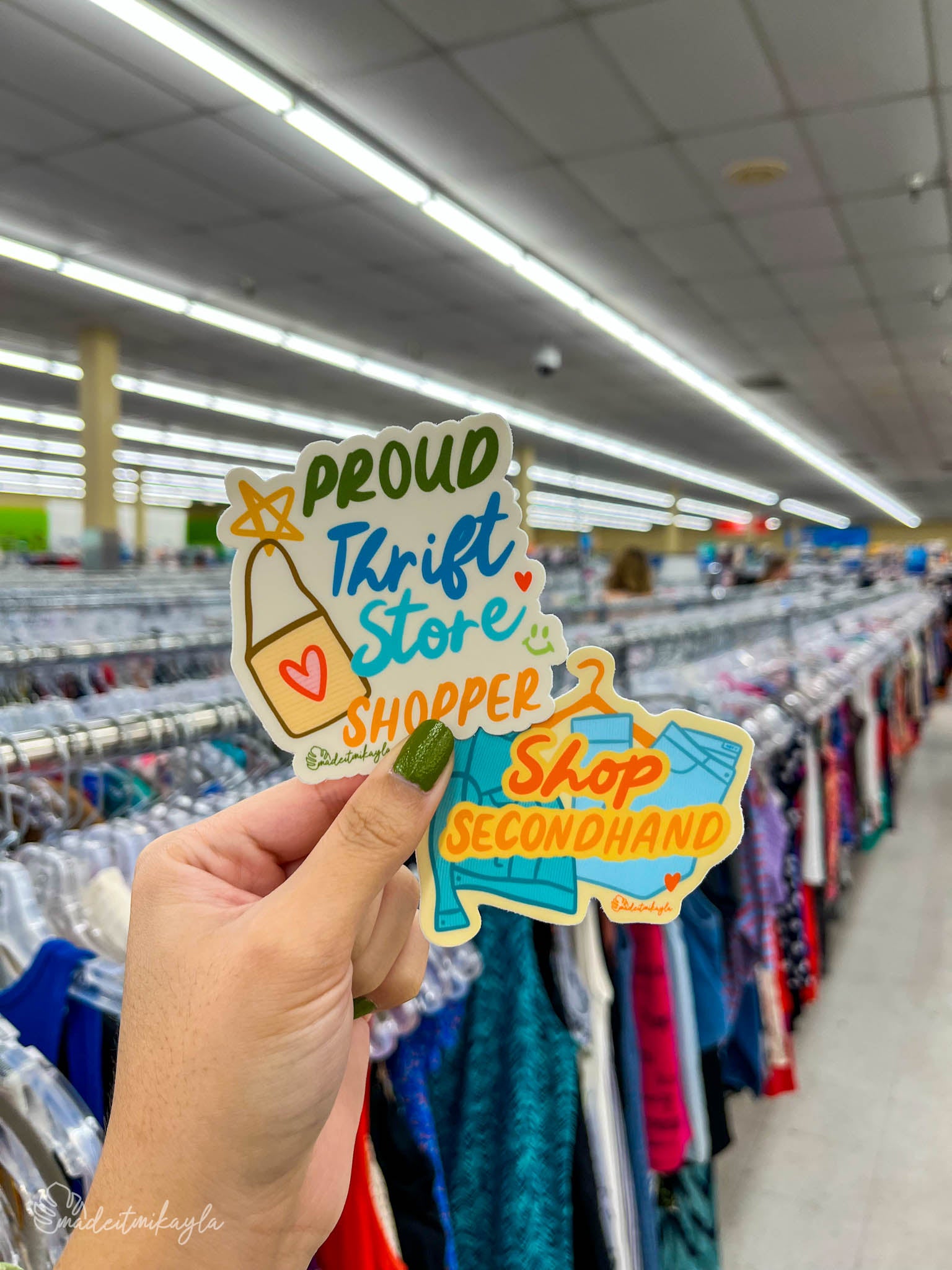 Hand holding a 'Proud Thrift Store Shopper' sticker and 'Shop Secondhand' sticker in a clothing thrift store.